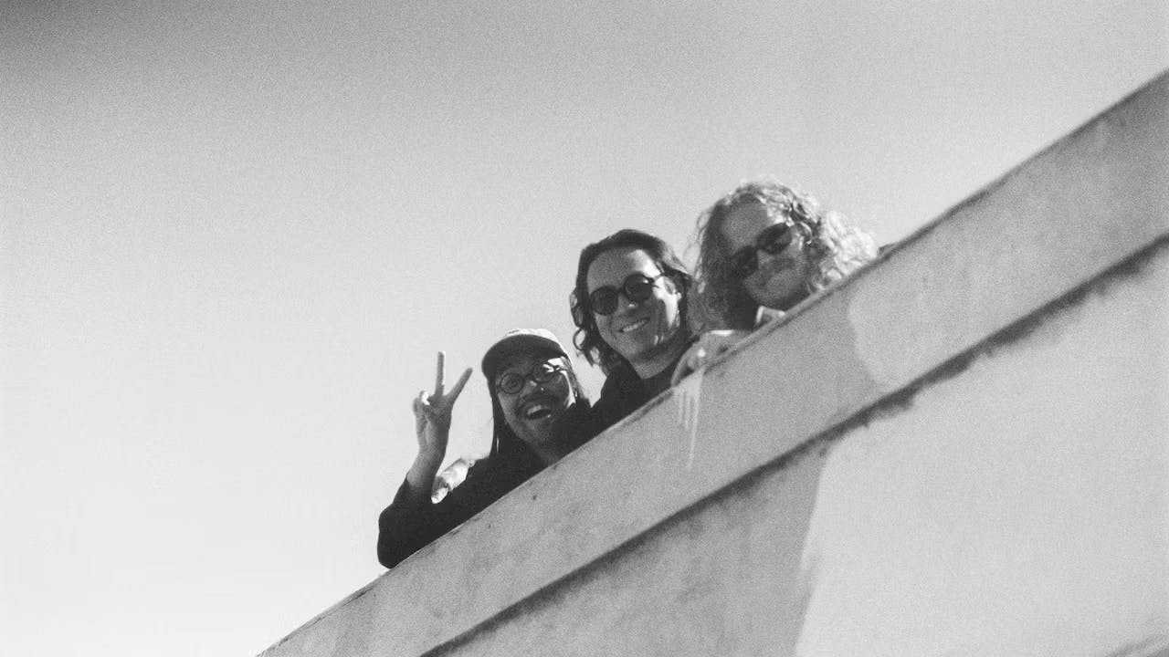 A black-and-white photo of Dave Harrington, Max Jaffe, and Patrick Shiorishi smiling from a parking garage rooftop