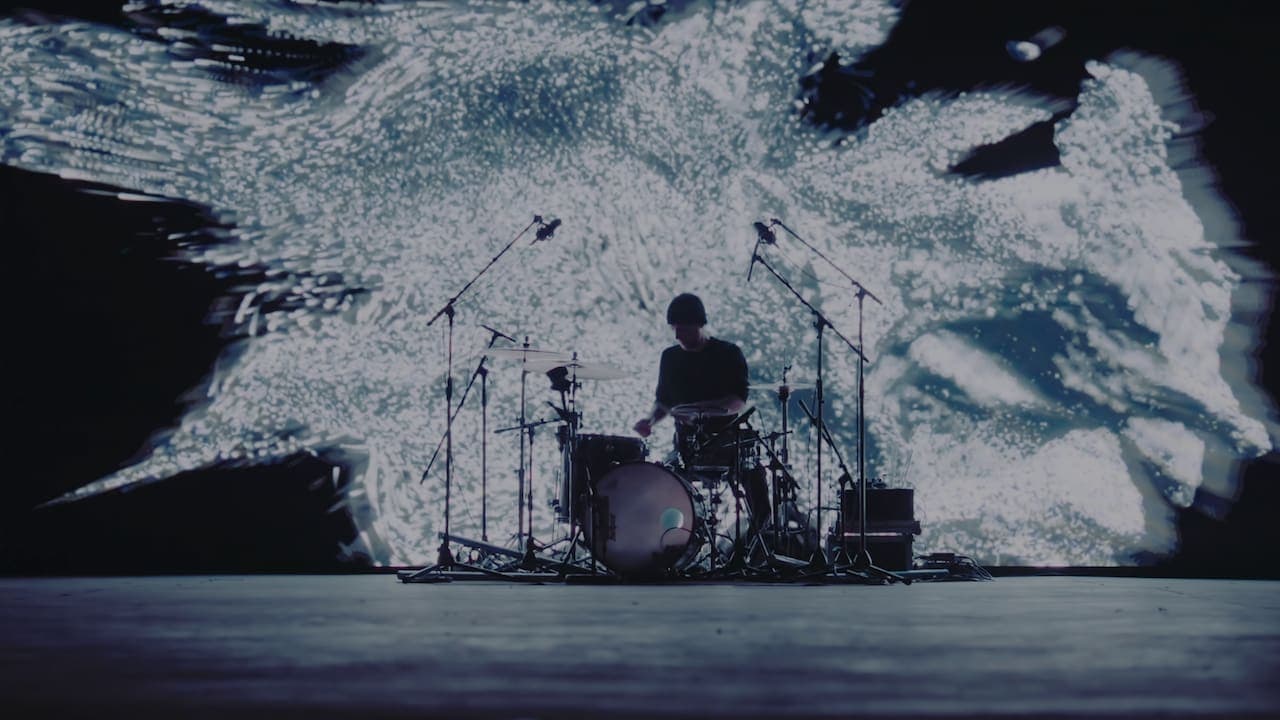 A photo of Khompa at the drum set in front of a large screen with a black and white 3D texture on it.