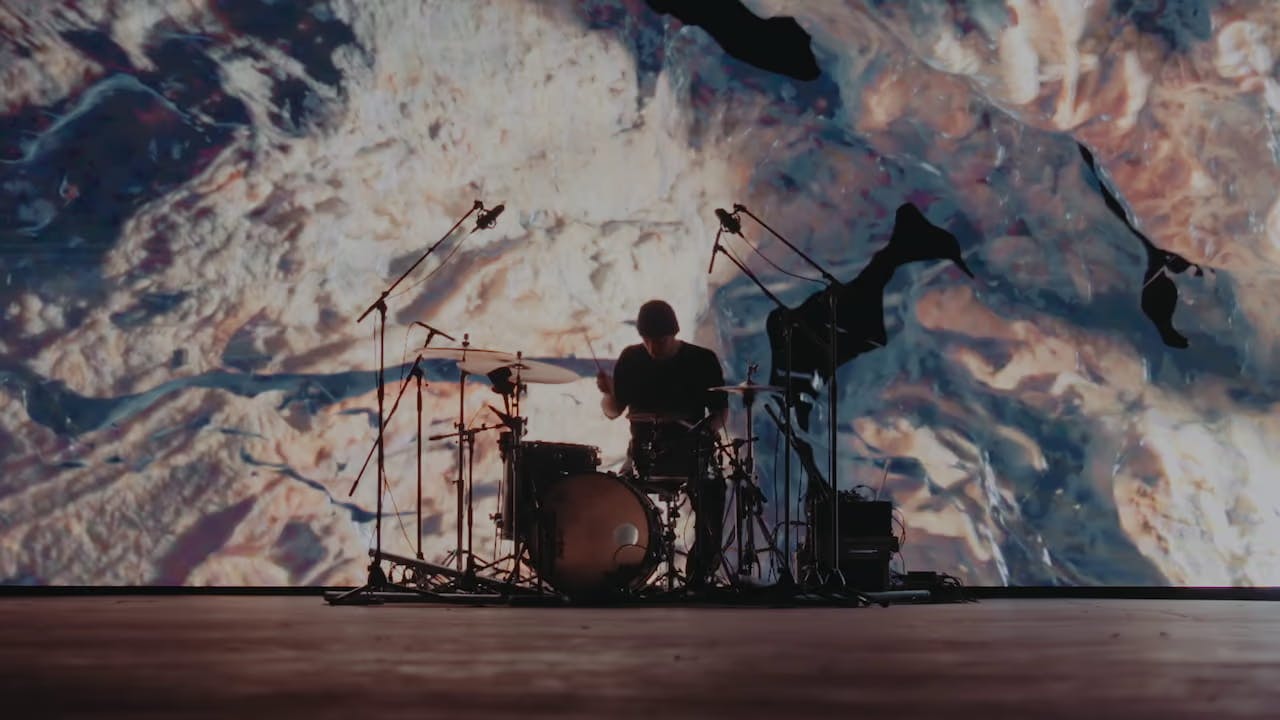 A photo of Khompa at the drum set in front of a large screen with a black and white 3D texture on it.