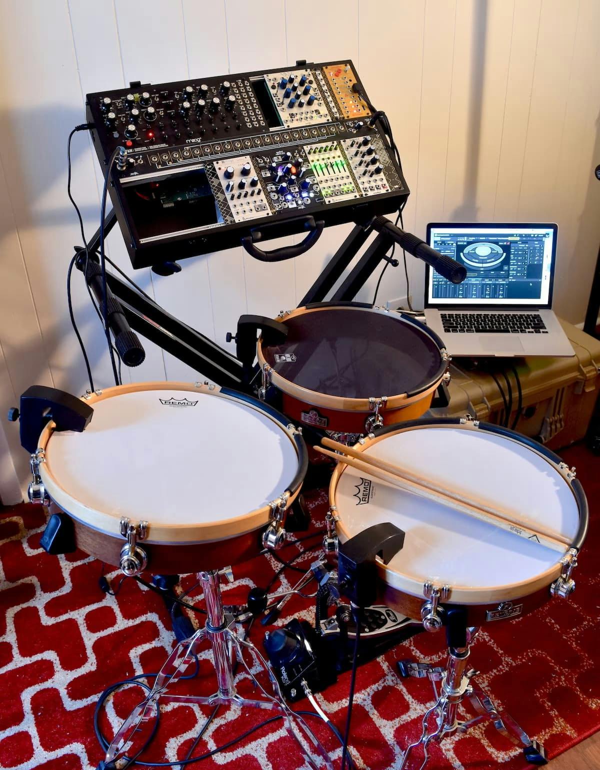 Three very shallow drums outfitted with Sunhouse sensors and arranged in a triangle, a Eurorack style synth filled with modules is in the background behind and above the drums, as is a laptop with Sensory Percussion software visible onscreen