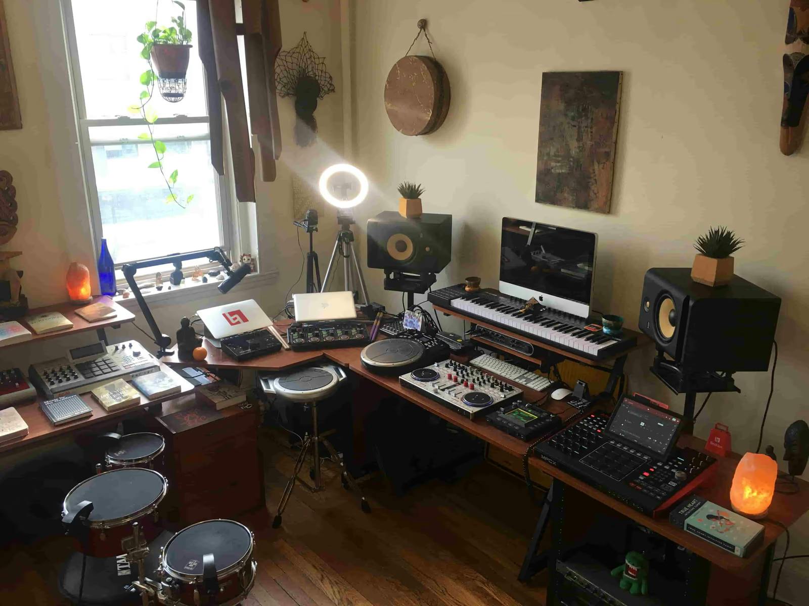 An overhead shot of Val's well lit studio showing all of the technology listed below (and more) with Afro-Hatian art on the walls, a hanging plant in the window, and one small potted plant on each studio speaker