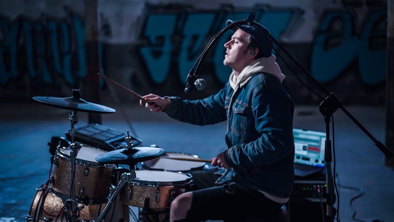 A side profile photo of Chordsplitter playing drums with a graffiti wall in the background.