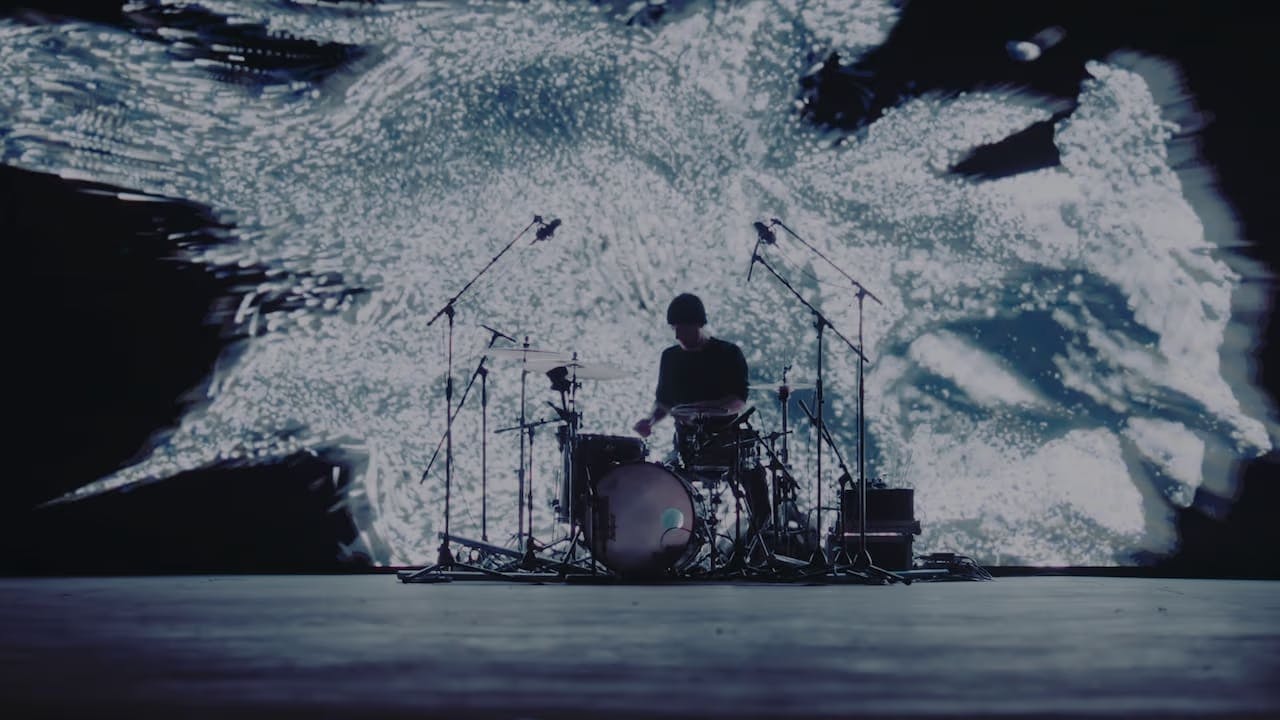 A photo of Khompa at the drum set in front of a large screen with a black and white 3D texture on it