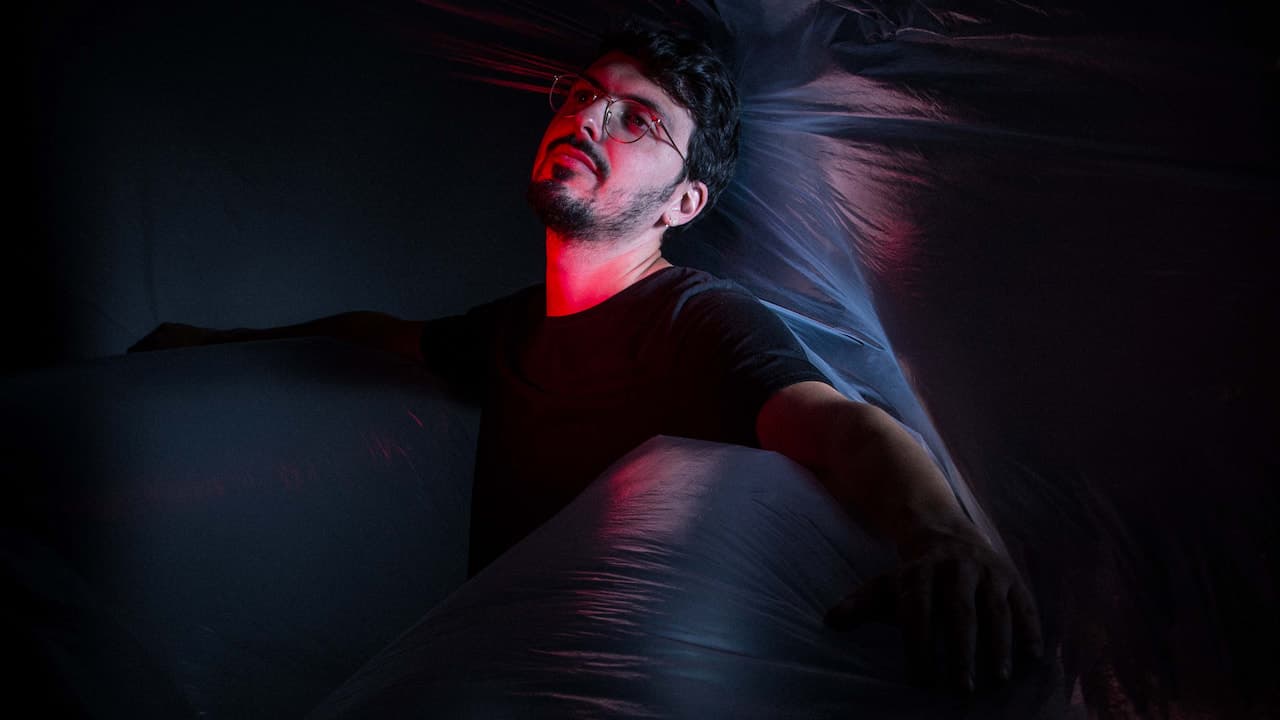 Antoine Pierre looking off into the distance with red lighting and a dark blue sheet blowing behind him