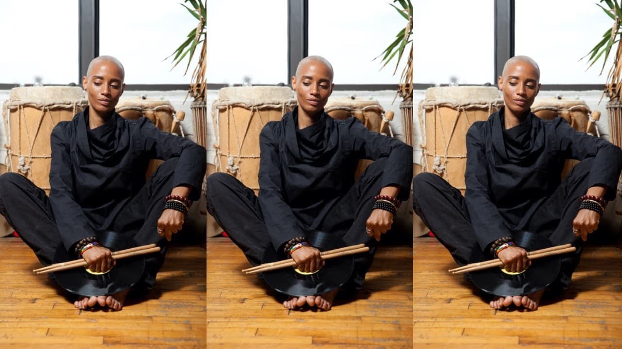 A photo of Val Jeanty seated in a butterfly pose on the floor in front of hand drums. She is dressed in all black and has drumsticks in her right hand, which is placed over a viynl record resting on her feet. Her left forearm is resting on her knee with her hand clenched in a fist.
